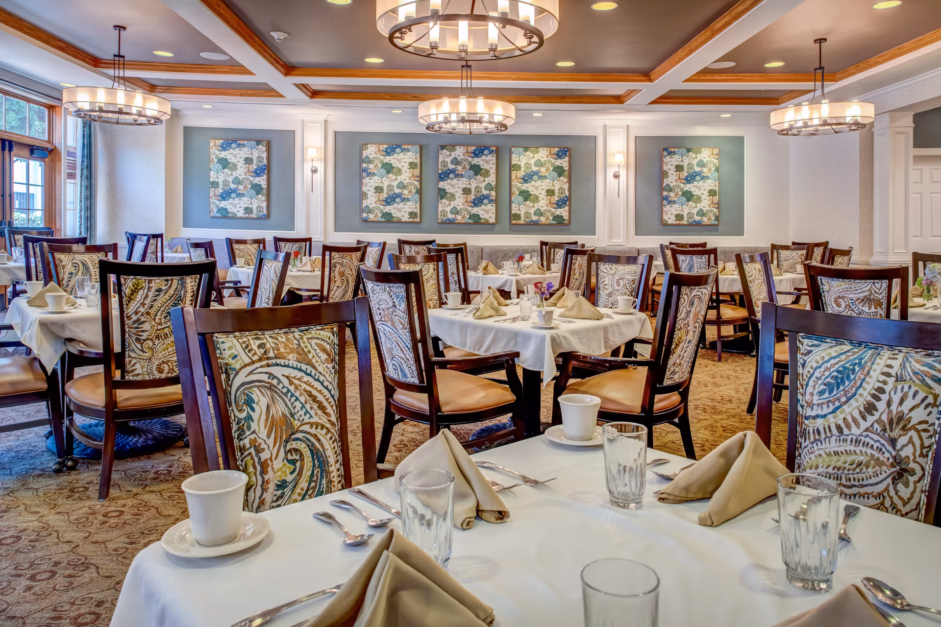 Bright, elegant dining room with multiple set tables, patterned chairs, chandeliers, and place settings.