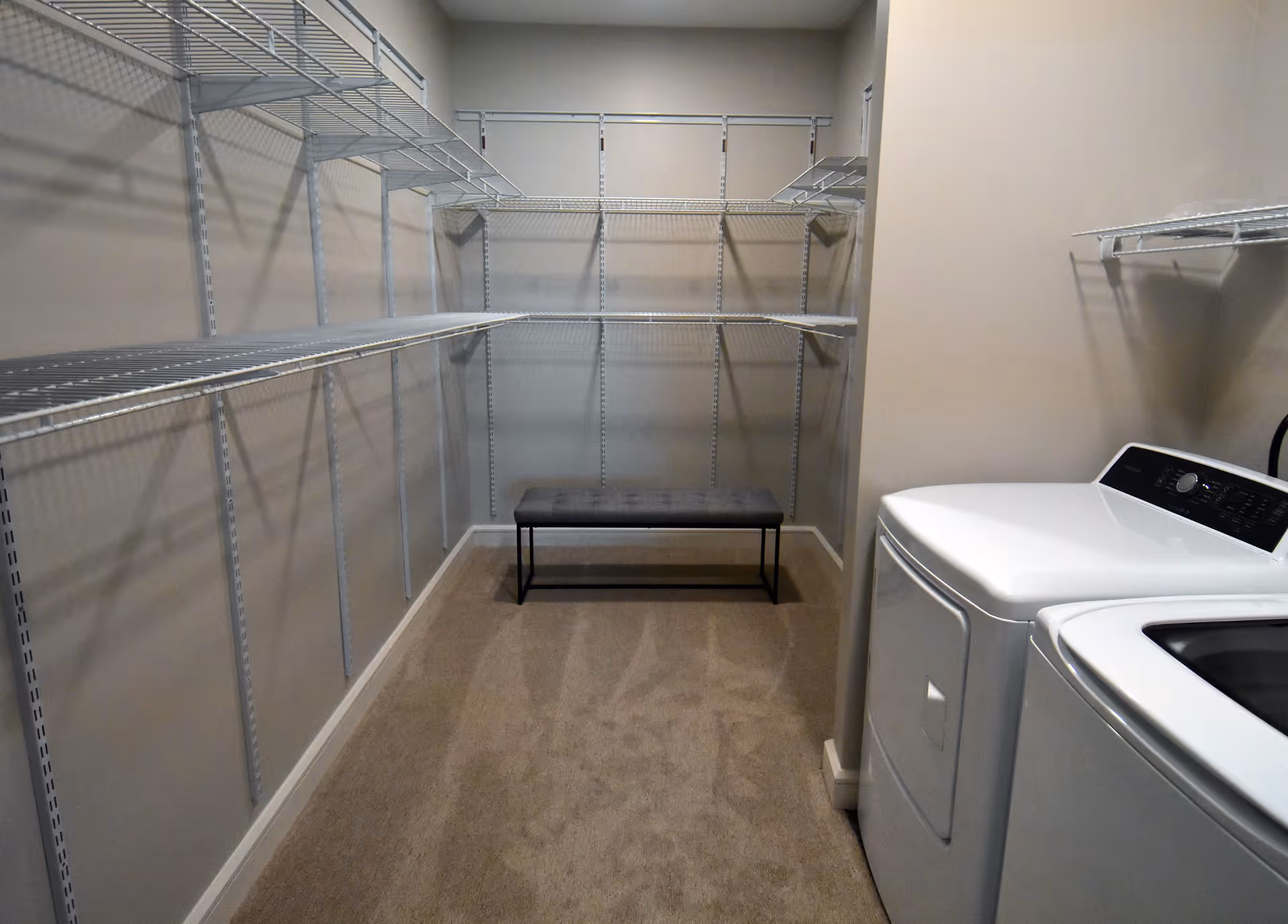 A laundry room with a white washing machine and dryer on the right side, beige carpeted floor, beige walls, multiple white wire shelves mounted on the walls, and a small gray bench at the far end of the room.