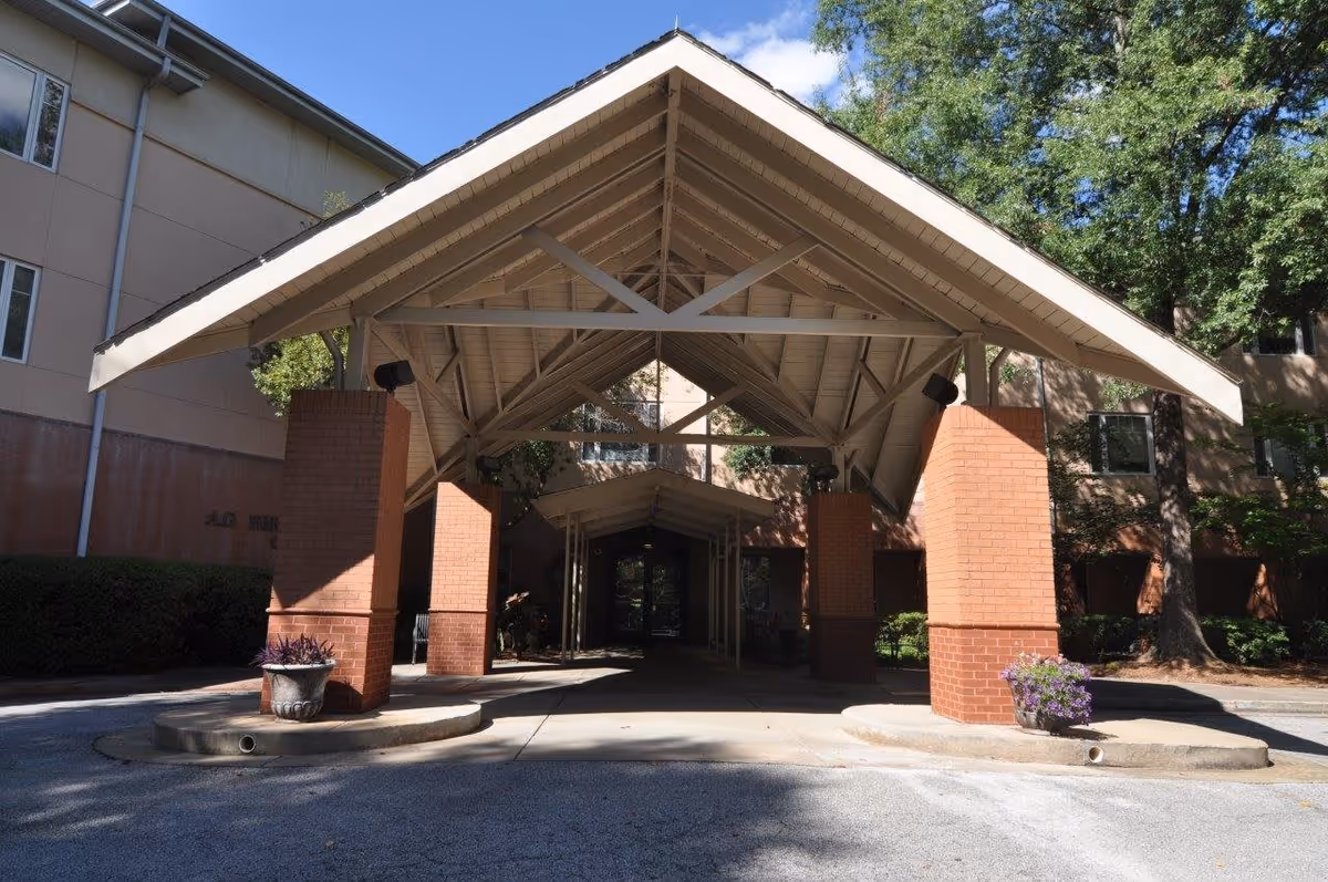 Entrance to a building with a large covered driveway supported by brick pillars, surrounded by trees and shrubs under a clear blue sky.