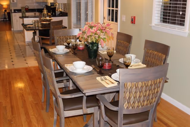 A dining room with a long wooden table set for six, upholstered chairs, a floral centerpiece, and a view into the adjacent kitchen.