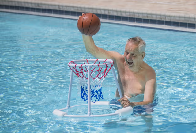 An elderly man in a swimming pool holding a basketball above a small floating basketball hoop, appearing joyful and engaged in a water activity.