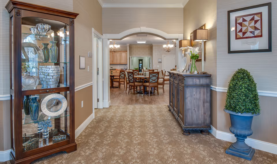 Carpeted entry hallway in a senior living facility leading into a dining area with wooden chairs, a glass display cabinet, and decorative furniture and plants.