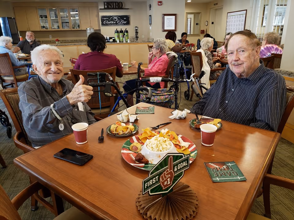Two elderly men sit at a table with snacks in a senior living dining area, one giving a thumbs-up while other residents eat in the background.