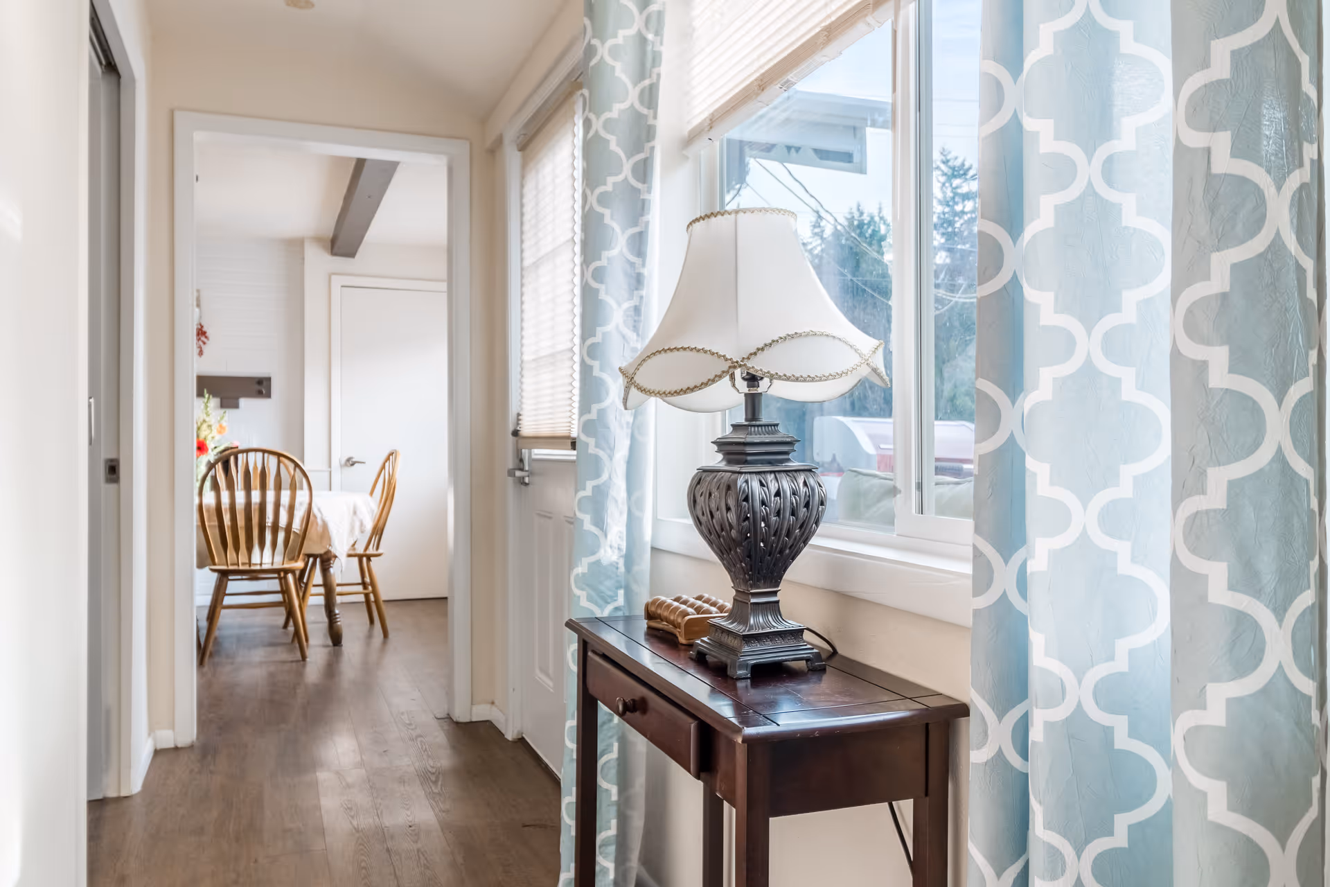 View down a hallway with a wooden side table holding a decorative lamp next to a window with blue and white patterned curtains. At the end of the hallway, a dining area is visible with a wooden table and chairs.