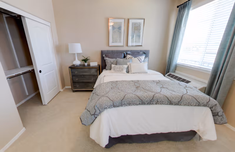 A neatly made bed with multiple pillows and a patterned comforter in a bedroom. Next to the bed is a dark wooden nightstand with a white lamp and a small plant. Two framed artworks hang above the bed. There is a window with light blue curtains on the right side, and an open closet with sliding doors on the left.