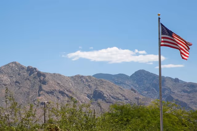 An American flag on a pole waving above desert shrubs with rocky mountains and a blue sky in the background.