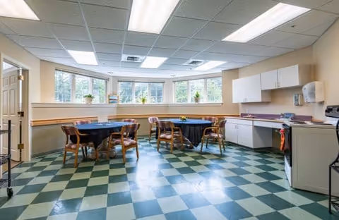 Bright communal dining/activity room with round tables and chairs, a kitchenette, and large bay windows over a green checkered floor.