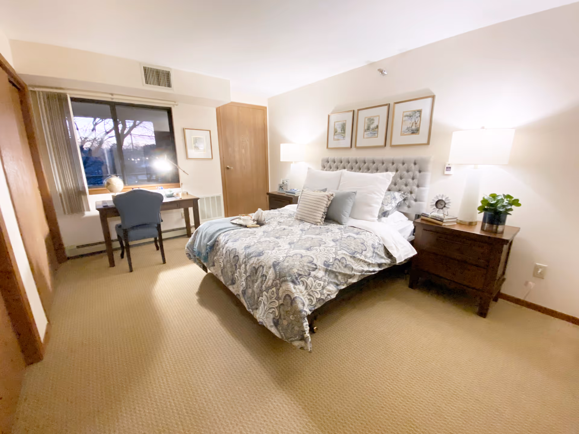 A cozy bedroom with a large bed featuring a tufted headboard and patterned bedding. There are two wooden nightstands on either side of the bed, each with a lamp and decorative items. A wooden desk with a blue chair and a globe sits by the window, which has vertical blinds partially open to reveal trees outside. The room has beige carpet and light-colored walls with three framed pictures above the bed.