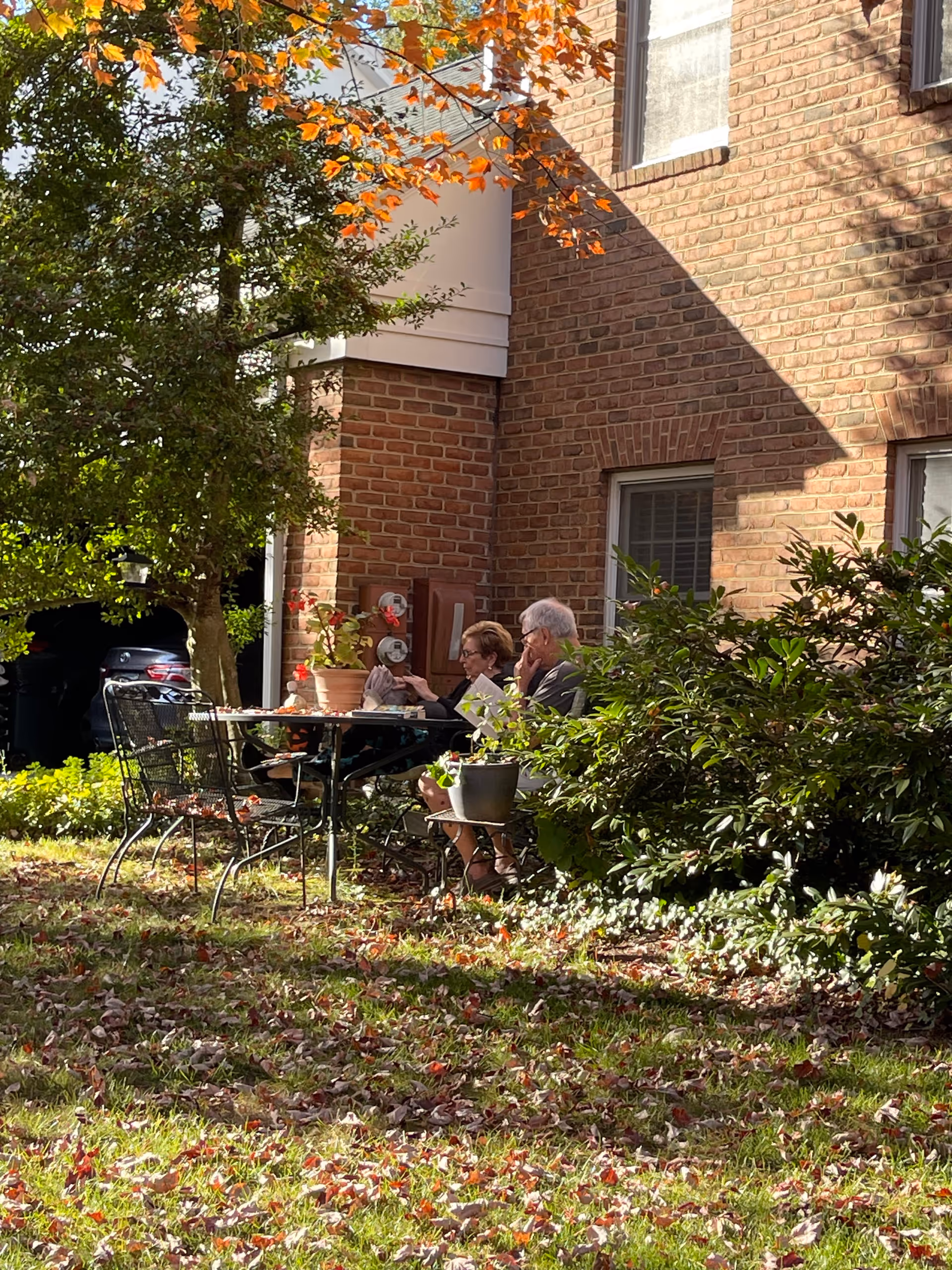 Two elderly people sitting at a metal outdoor table with chairs in a garden area next to a brick building. The table has potted plants on it, and there are trees and bushes around. The ground is covered with fallen leaves.