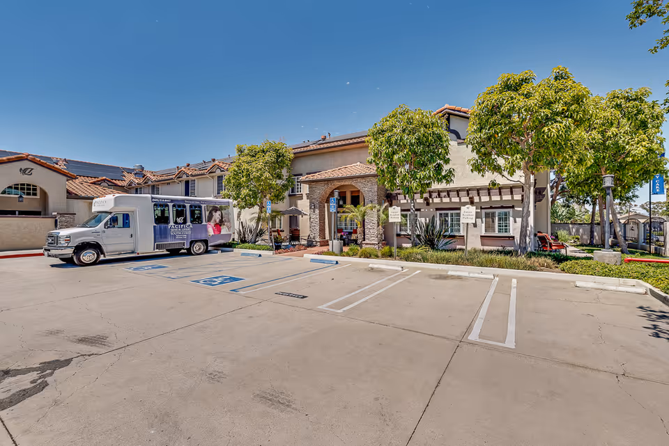 Exterior view of Pacifica Senior Living South Coast building with a parking lot in front. There is a shuttle bus parked in a handicapped parking space, several trees, and clear blue sky.