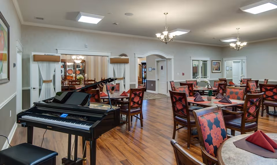 A spacious dining room in a senior living facility featuring multiple tables with red and purple floral-patterned chairs. Each table is set with folded napkins and small flower arrangements. A black grand piano is positioned near the left side of the room. The room has wooden flooring, soft lighting from chandeliers, and light-colored walls with white trim. There are doorways and windows leading to other rooms in the background.
