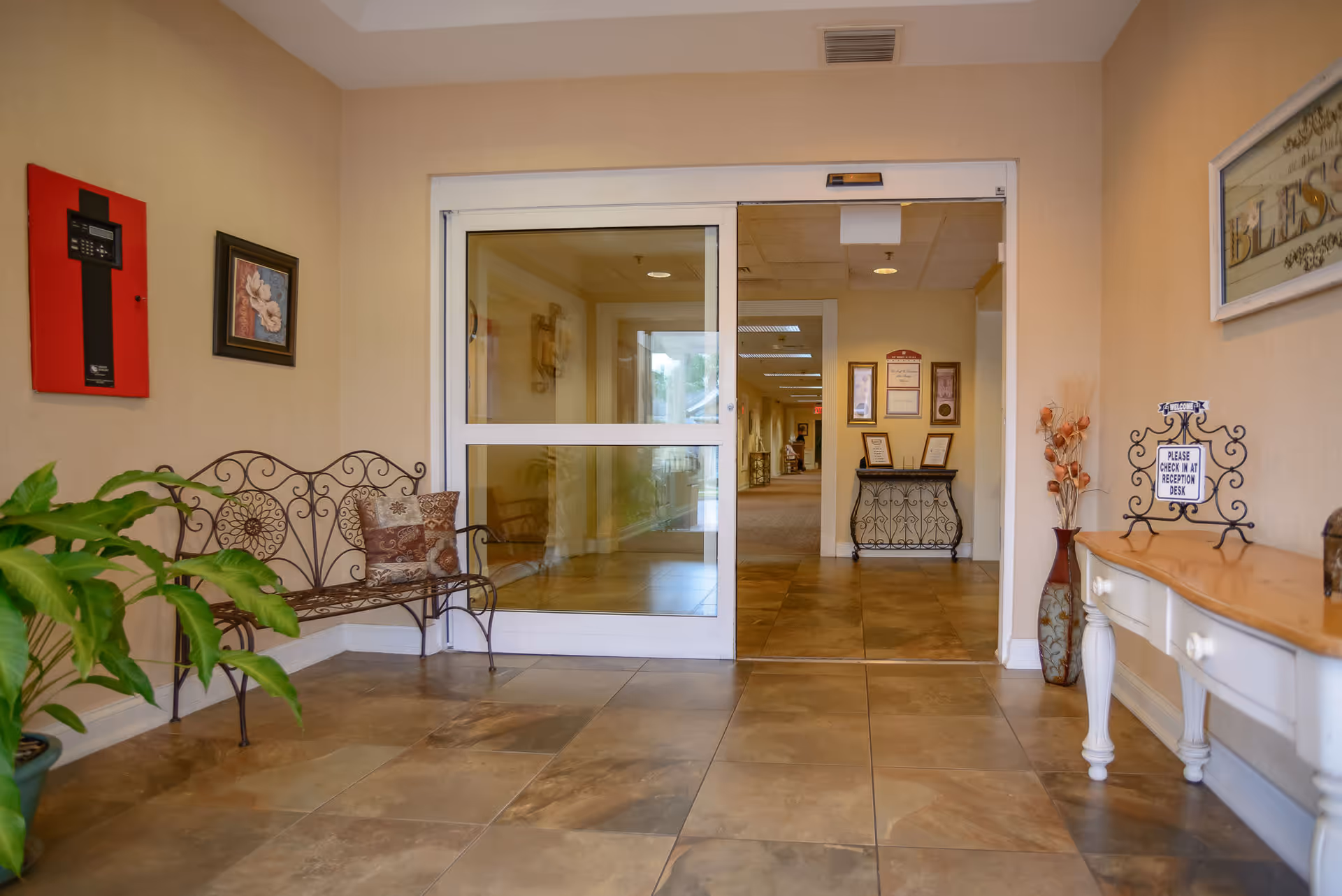 Entrance area of a senior living facility with a decorative metal bench with cushions on the left, a potted plant, and a white wooden console table on the right with a sign that reads 'Please check in at reception desk.' There is a sliding glass door leading to a hallway with framed certificates and a small decorative table visible inside.
