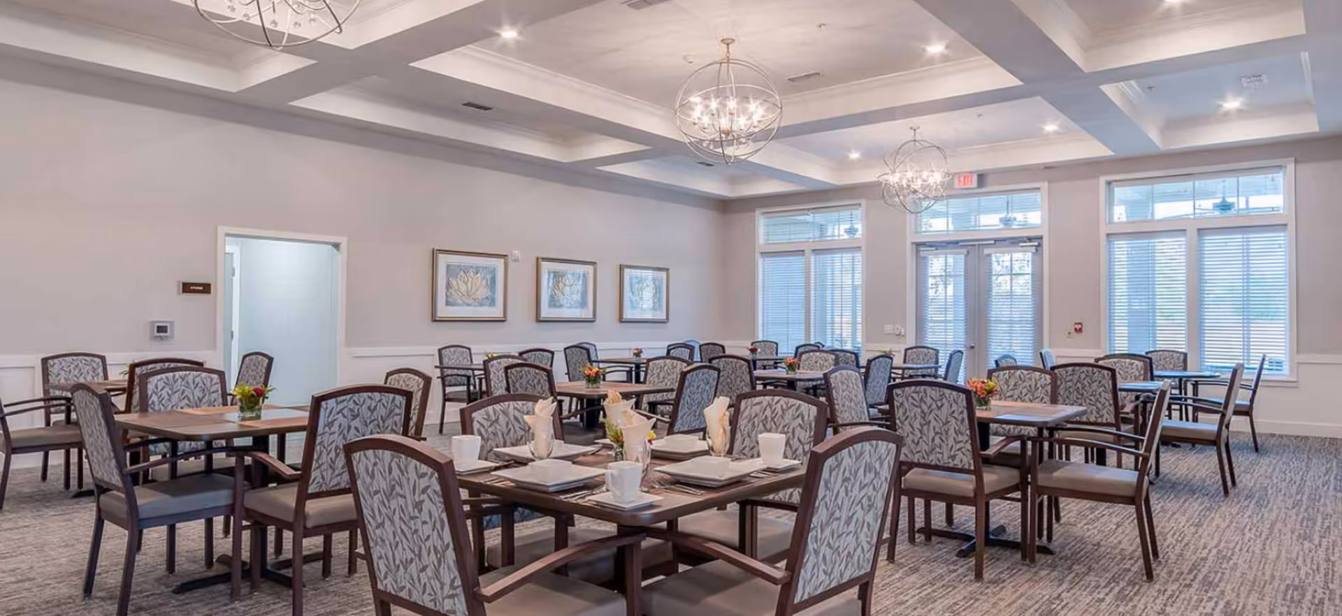 A spacious dining room with multiple tables and chairs arranged neatly. Each table is set with white plates, cups, and napkins, and small floral centerpieces. The room features large windows with blinds, three framed artworks on the wall, and two modern chandeliers hanging from a coffered ceiling.