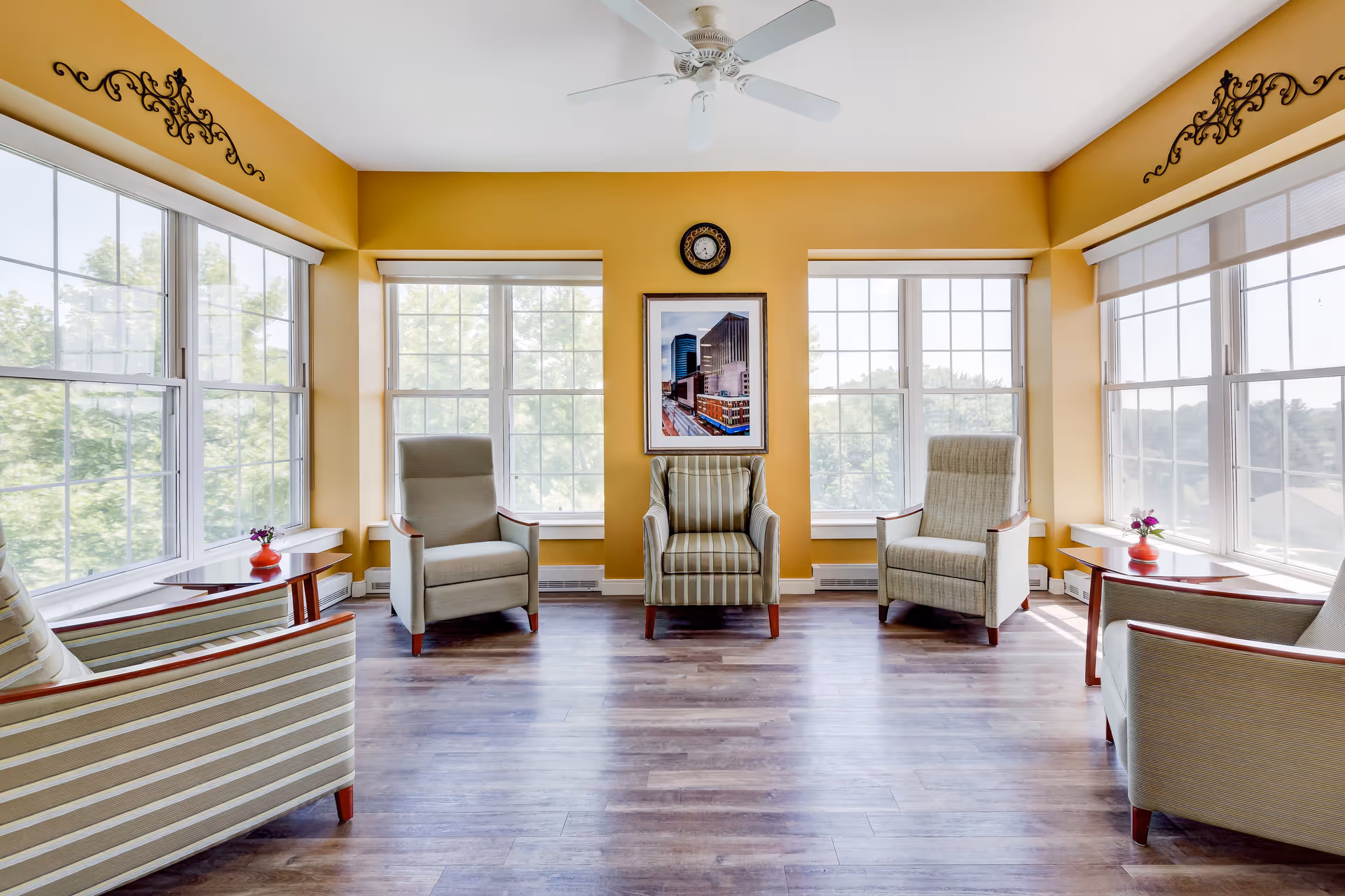 Sunny communal sitting room with multiple upholstered armchairs arranged by large windows and warm yellow walls.