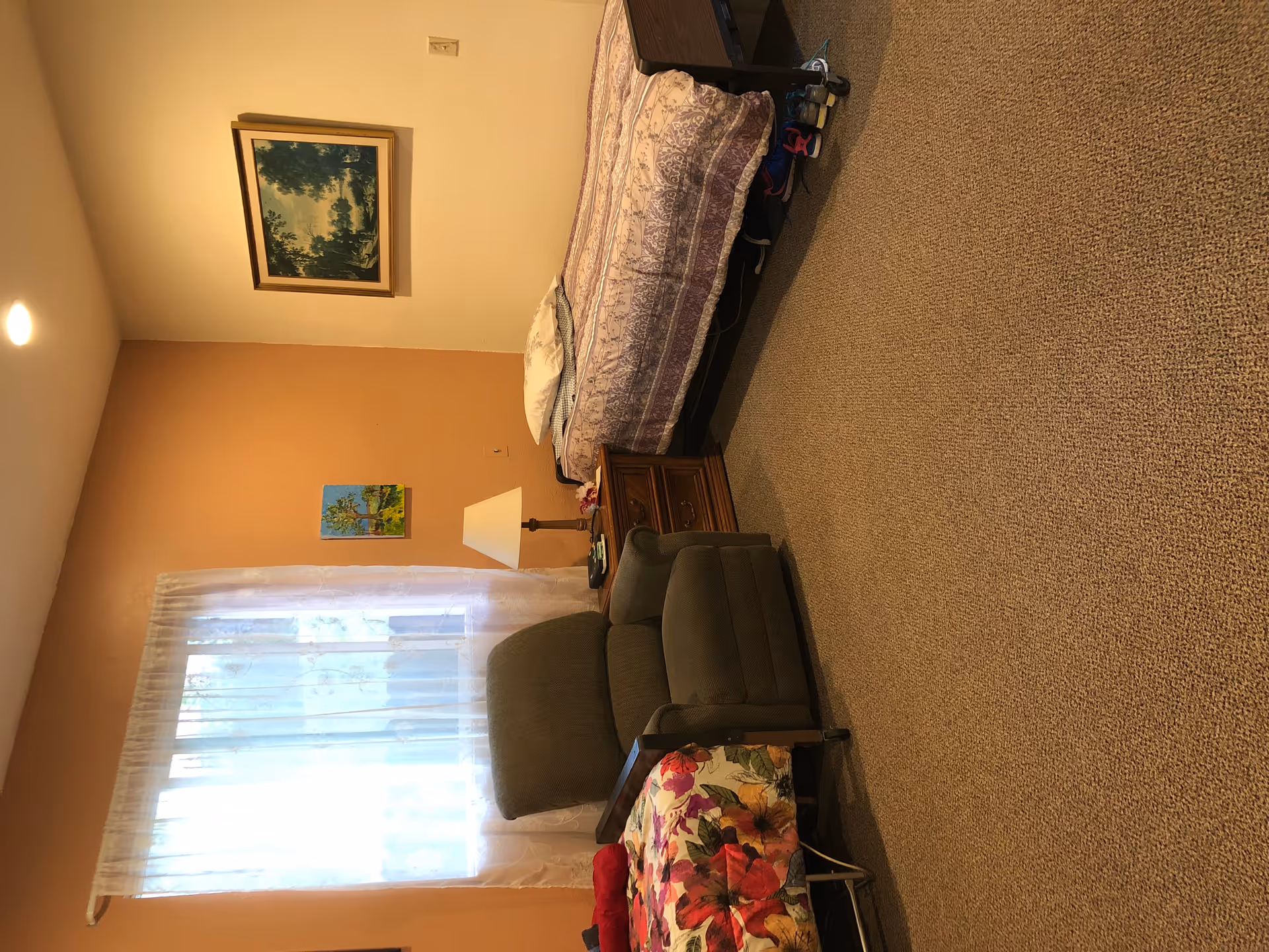 A cozy bedroom with two single beds, one with a floral bedspread and the other with a patterned purple and white bedspread. Between the beds is a wooden nightstand with a lamp and some small items. A comfortable armchair is positioned near a window with sheer white curtains. The walls are painted in two tones, one side orange and the other beige, and there are two framed paintings hanging on the walls.