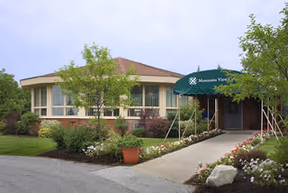 Exterior view of a single-story building with a green awning labeled 'Mountain View Center' over the entrance. The building is surrounded by landscaped greenery, including trees, shrubs, and flower beds along a paved walkway leading to the door.