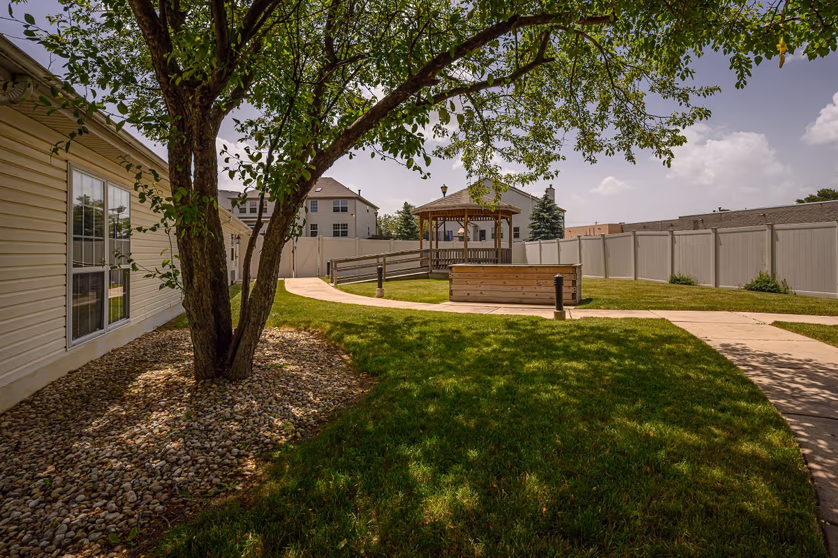 Outdoor area of Arden Courts - ProMedica Memory Care Community in Elk Grove featuring a large tree with green leaves, a grassy lawn, a paved walkway, a wooden planter box, and a gazebo in the background surrounded by a white fence.