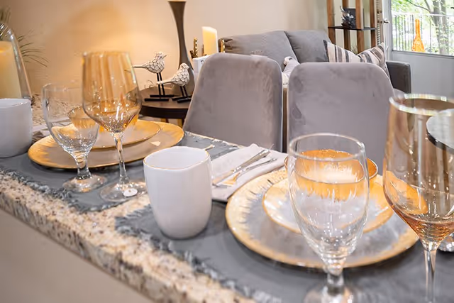 Close-up view of a dining counter set with gold-rimmed plates, clear and amber-colored wine glasses, white mugs, and silverware on a gray placemat. In the background, there are gray upholstered chairs and a living room area with a sofa and decorative items.
