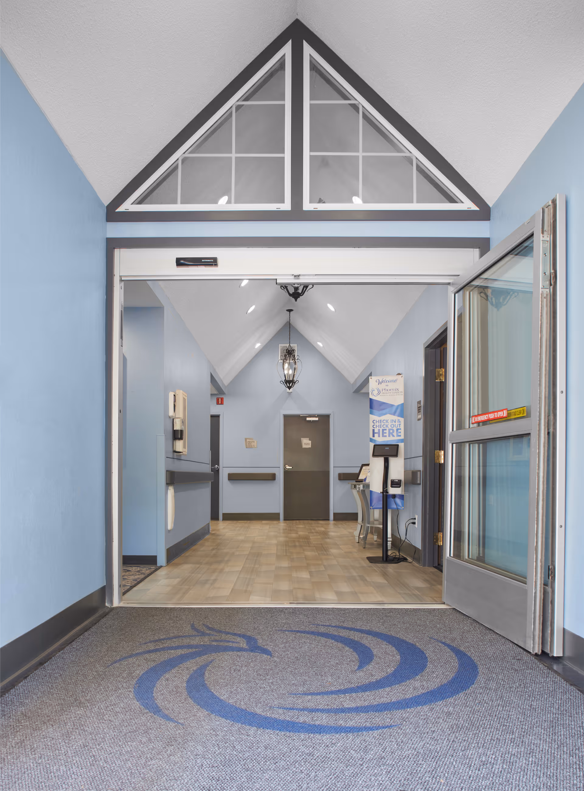 Entrance hallway of a facility with light blue walls, a gray door at the end, a hanging lantern light fixture, and a check-in kiosk with a sign that says 'Welcome Phoenix of Fairlawn, Check In or Check Out Here'. The floor has a light wood pattern and there is a gray mat with a blue swirl logo near the entrance.