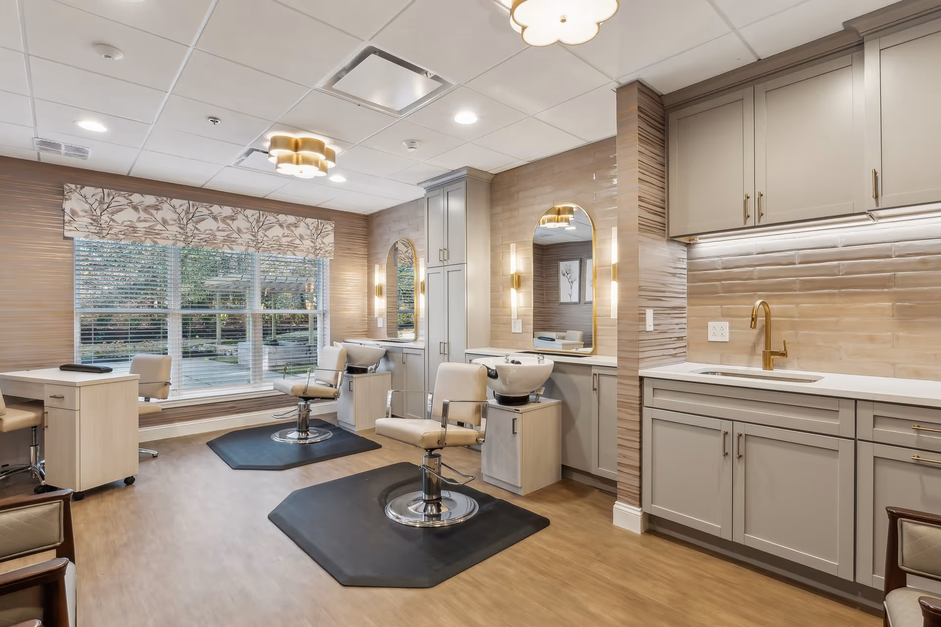 A bright and modern salon area in a senior living facility with two salon chairs in front of wash basins and mirrors. The room features light wood flooring, beige walls with textured tiles, large windows with floral valances, and cabinetry with a gold faucet and under-cabinet lighting.