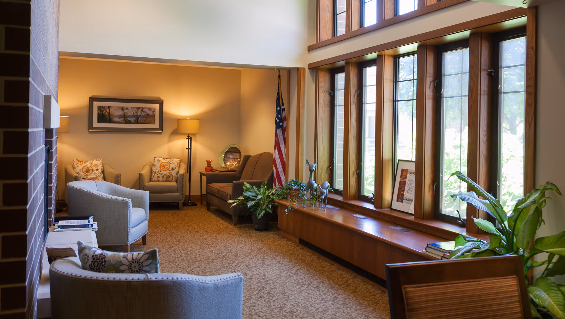 A cozy living room area with several upholstered chairs and a sofa arranged around a carpeted floor. There is a floor lamp and framed artwork on the wall. Large windows with wooden frames let in natural light, and various plants and decorative items are placed on a wooden ledge beneath the windows. An American flag stands in the corner.