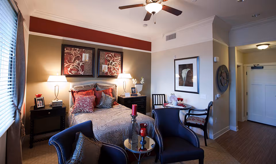 A cozy bedroom in a senior living facility featuring a bed with patterned bedding and multiple pillows, two bedside tables with lamps, framed artwork above the bed, two navy blue armchairs with a small round table between them, a small dining table with two chairs, a large wall clock, and a ceiling fan with light. The room has a window with blinds and curtains, and a hallway leading to a white door.