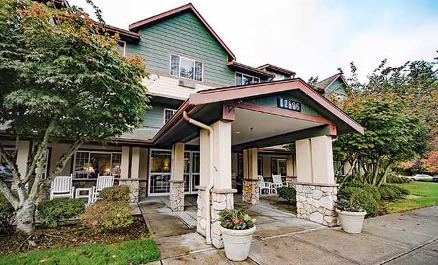 Front exterior view of Brookdale Arbor Place senior living facility showing a covered entrance with stone pillars, white rocking chairs on the porch, green and beige siding, and surrounding trees and shrubs.
