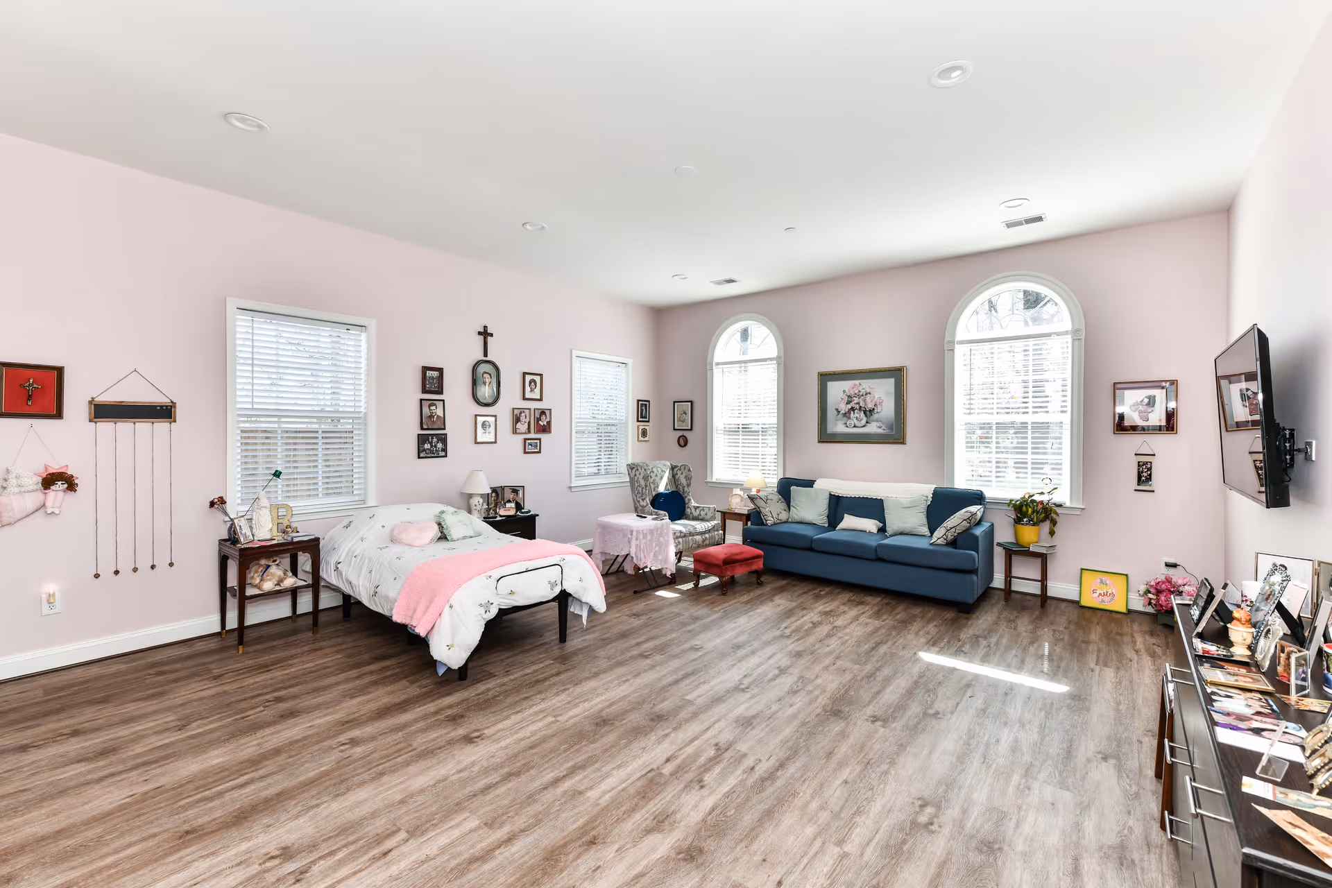 A spacious, well-lit bedroom with light pink walls and wood flooring. The room features a single bed with white bedding and a pink throw, a blue sofa with cushions, a patterned armchair with a small round table covered by a pink cloth, and several framed pictures and decorations on the walls. There are three windows with white blinds allowing natural light to fill the room. A flat-screen TV is mounted on the wall opposite the bed, and a dresser with various framed photos and items is below the TV.