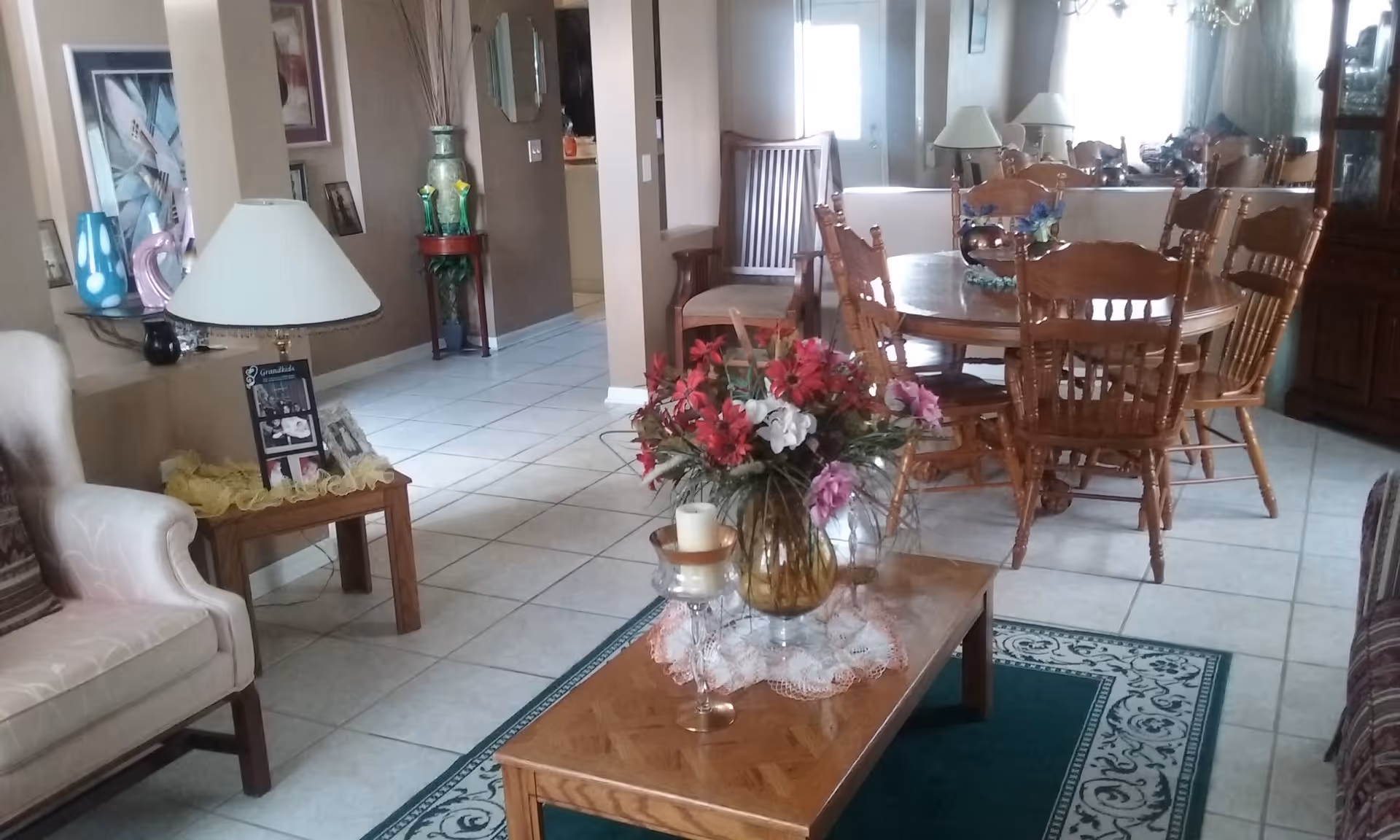 Interior view of a living and dining area with a wooden dining table and chairs, a coffee table with a floral arrangement and candle, a beige armchair, a side table with a lamp and photo frames, and decorative items on the walls and shelves.