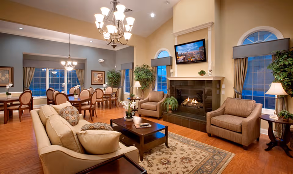 A cozy and well-lit common area in a senior living facility featuring a beige sofa with patterned cushions, two armchairs, a wooden coffee table on a floral rug, a fireplace with a mounted TV above it, and a dining area with multiple tables and chairs near large windows with curtains.