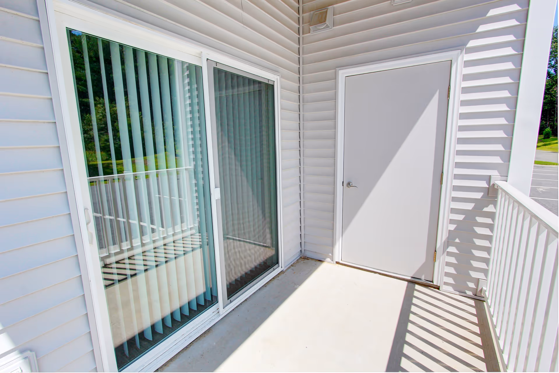Small outdoor balcony with a sliding glass door with vertical blinds, a white entry door, and a railing casting striped shadows.