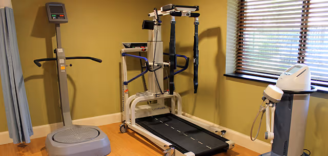 A rehabilitation room with exercise and therapy equipment including a treadmill with harness support, a vibration plate machine, and another medical device, all set against a green wall with a window covered by blinds.