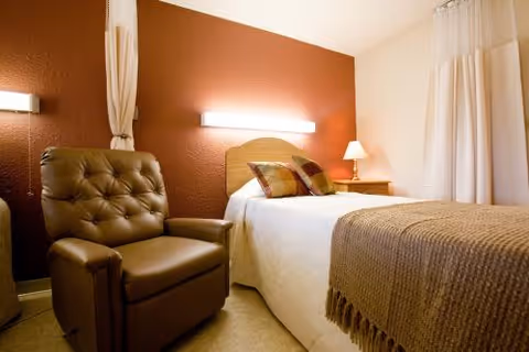 A cozy bedroom with a single bed covered in white sheets and a brown throw blanket, two pillows, a wooden headboard, a nightstand with a lamp, a brown leather recliner chair, and white curtains on the window. The walls are painted a warm reddish-brown color.