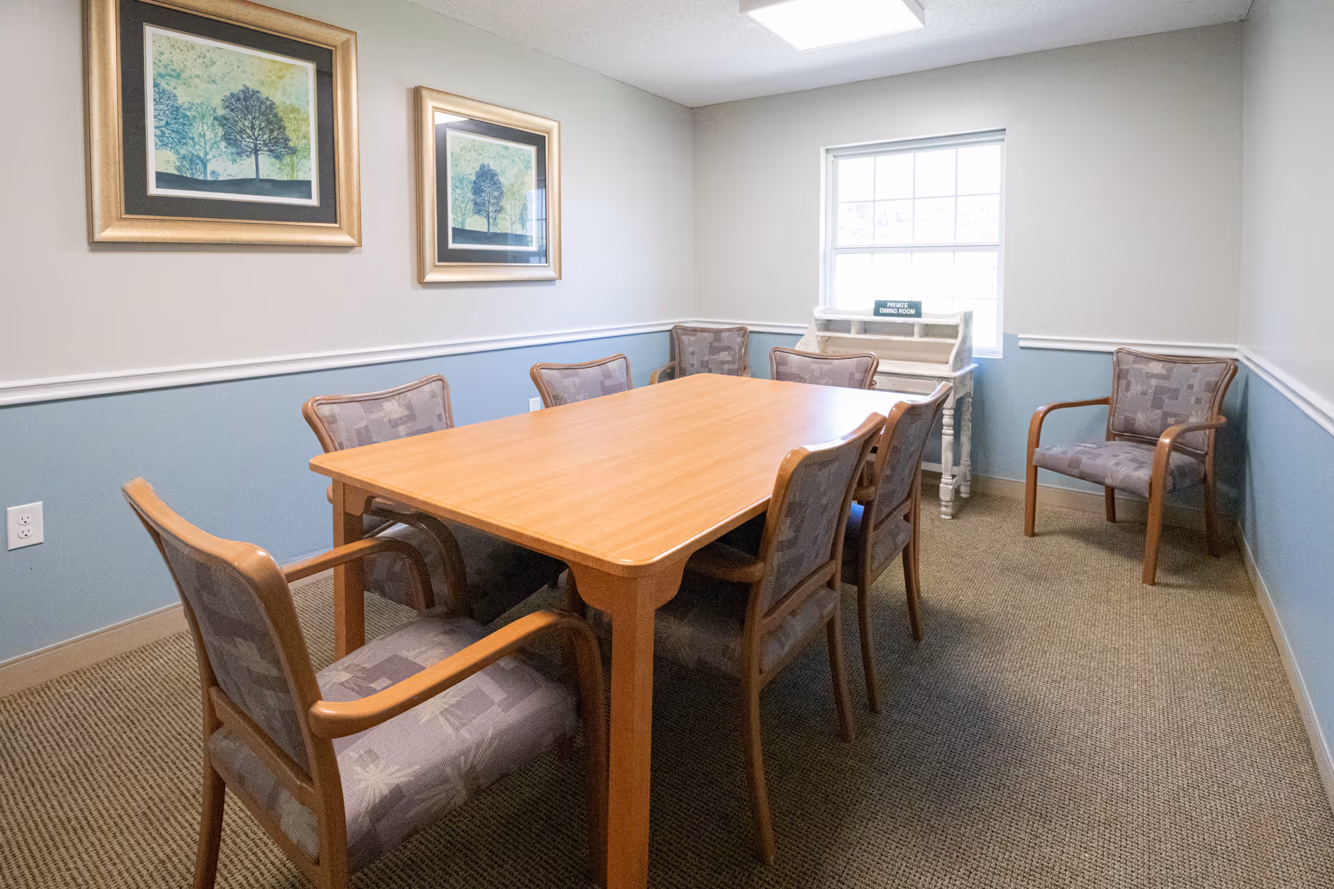 A small dining room with a rectangular wooden table surrounded by six cushioned chairs with wooden frames. The walls are painted light gray on the upper half and blue on the lower half, with white trim in between. Two framed pictures of trees hang on the left wall. A window on the far wall lets in natural light, and below it is a small white wooden cabinet with a sign that reads 'PRIVATE DINING ROOM'.