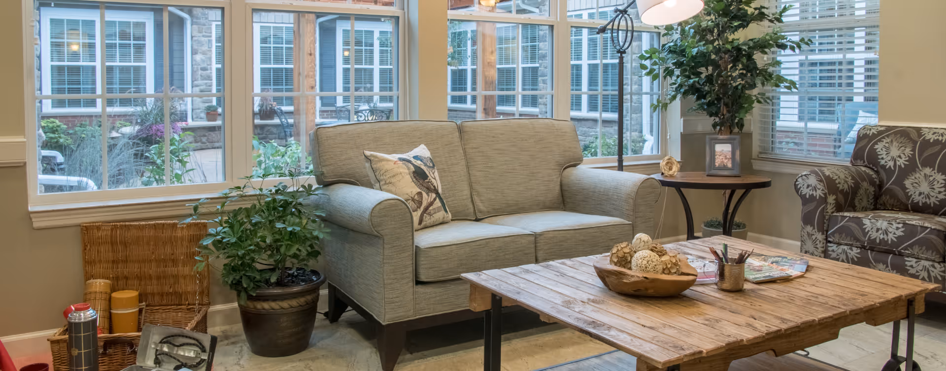 A cozy living room area with a beige loveseat adorned with a decorative pillow, a wooden coffee table with decorative items and magazines, a potted plant, a wicker basket, and a floral-patterned armchair. Large windows provide a view of an outdoor courtyard with additional seating and greenery.
