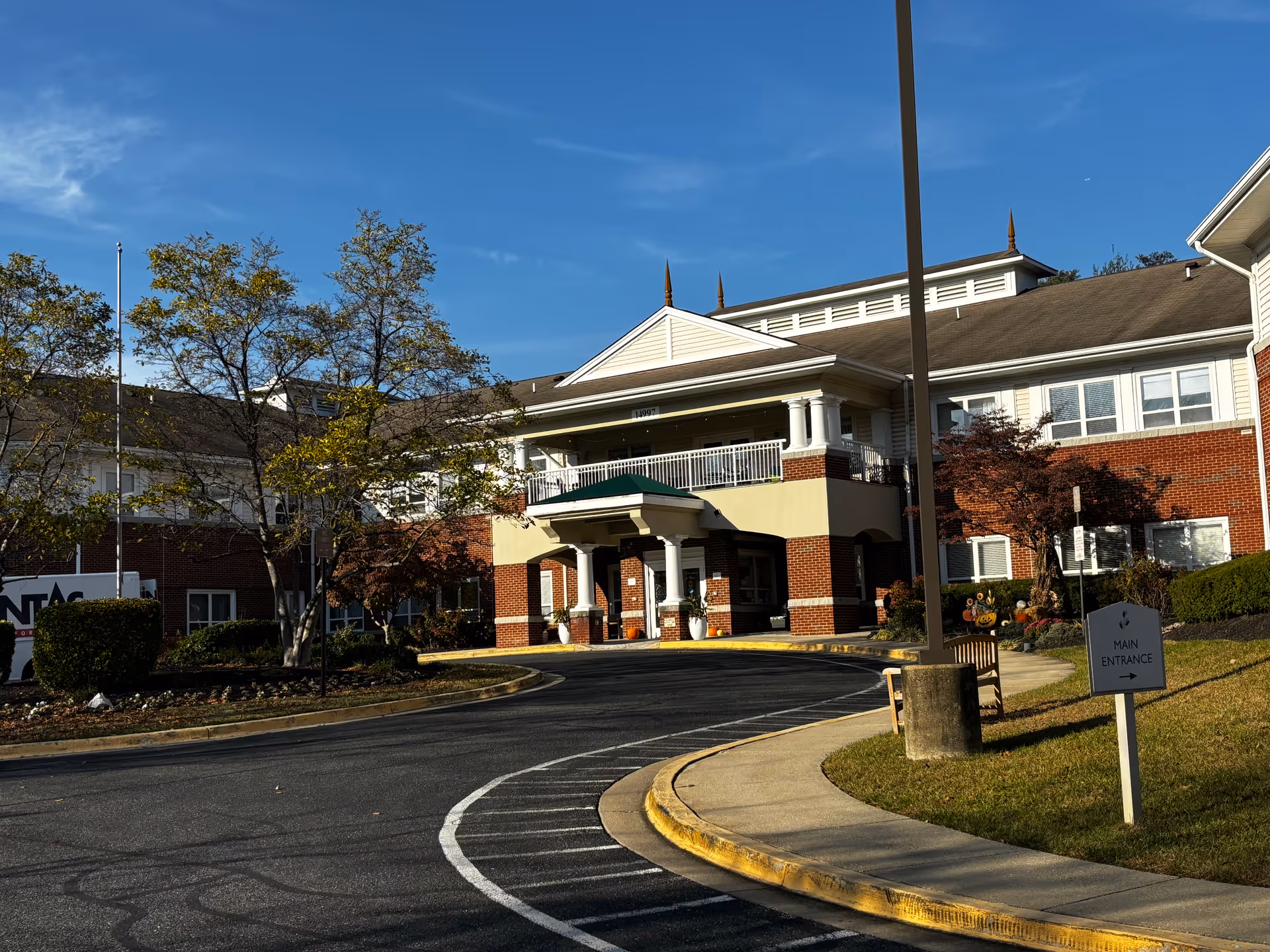Entrance and curved driveway of a brick-and-siding senior living building with a covered portico, landscaping, and a 'Main Entrance' sign.