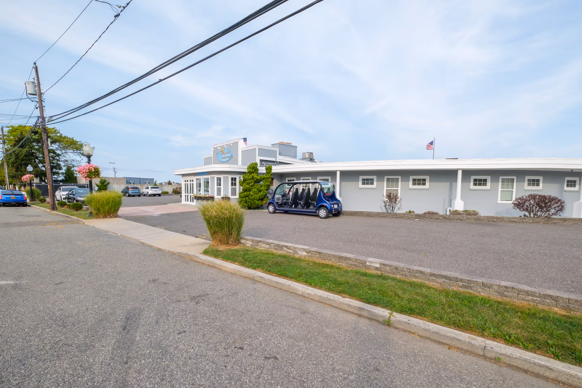 Exterior view of Babylon Beach House facility with a parking area, a blue golf cart, and a clear sky. The building is light gray with white trim and has an American flag on the roof.
