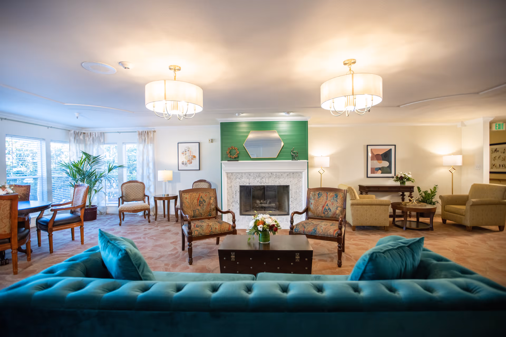A spacious and well-lit senior living facility common area with a teal tufted sofa in the foreground, two patterned armchairs facing a white fireplace with a green accent wall and a hexagonal mirror above it. The room also features additional seating including chairs and small tables, decorative plants, framed artwork on the walls, and two ceiling light fixtures.
