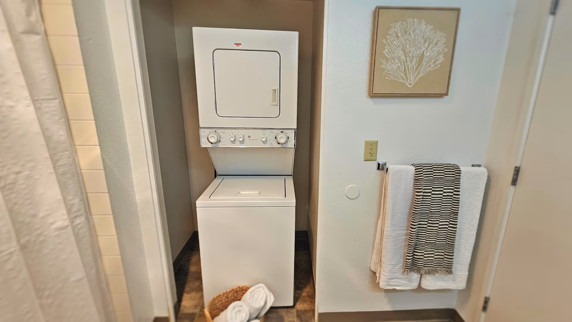 A small laundry area with a stacked washer and dryer unit. To the right, there is a towel rack mounted on the wall holding two white towels and one patterned towel. Above the towel rack, there is a framed artwork of a coral design. On the floor in front of the washer and dryer, there is a basket with rolled white towels. The walls are light-colored and the floor has a dark tile pattern.