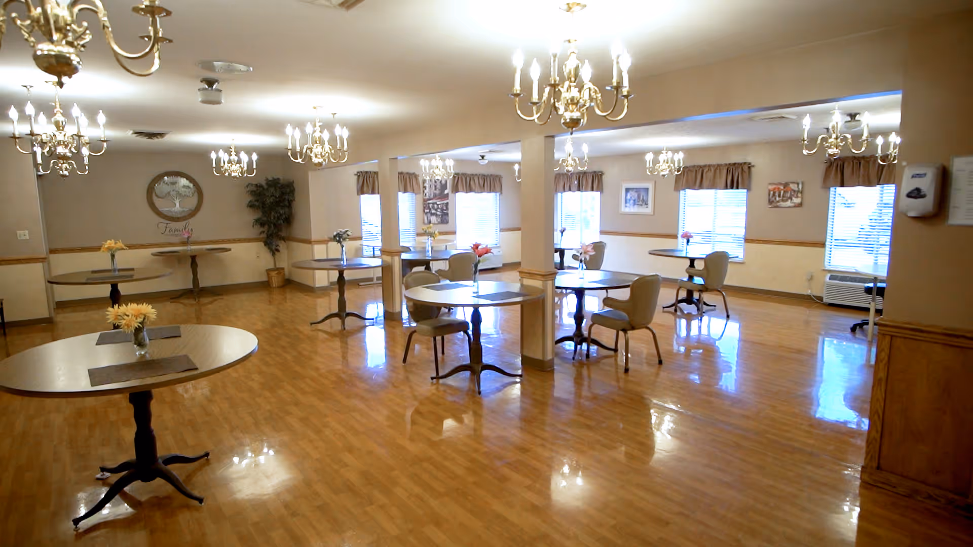 A spacious dining room with multiple round wooden tables, each adorned with a small vase of flowers. The room has polished wooden floors, several chandeliers hanging from the ceiling, and windows with beige curtains letting in natural light. There are a few chairs around some tables and framed pictures on the walls.