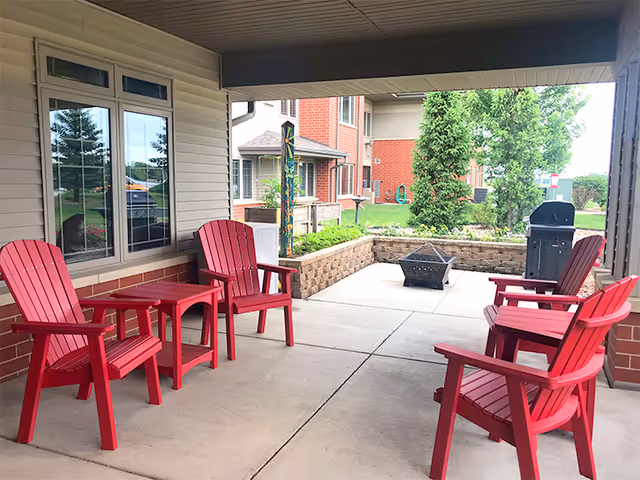 Covered outdoor patio area with four red Adirondack chairs and two small red tables arranged on a concrete floor. In the background, there is a fire pit, a barbecue grill, and a raised garden bed with greenery. The patio is adjacent to a building with beige siding and windows, and there are trees and other buildings visible beyond the patio.