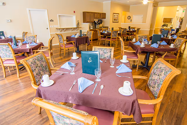 Bright dining room with multiple tables set with tablecloths, place settings, glassware, and floral-upholstered chairs.