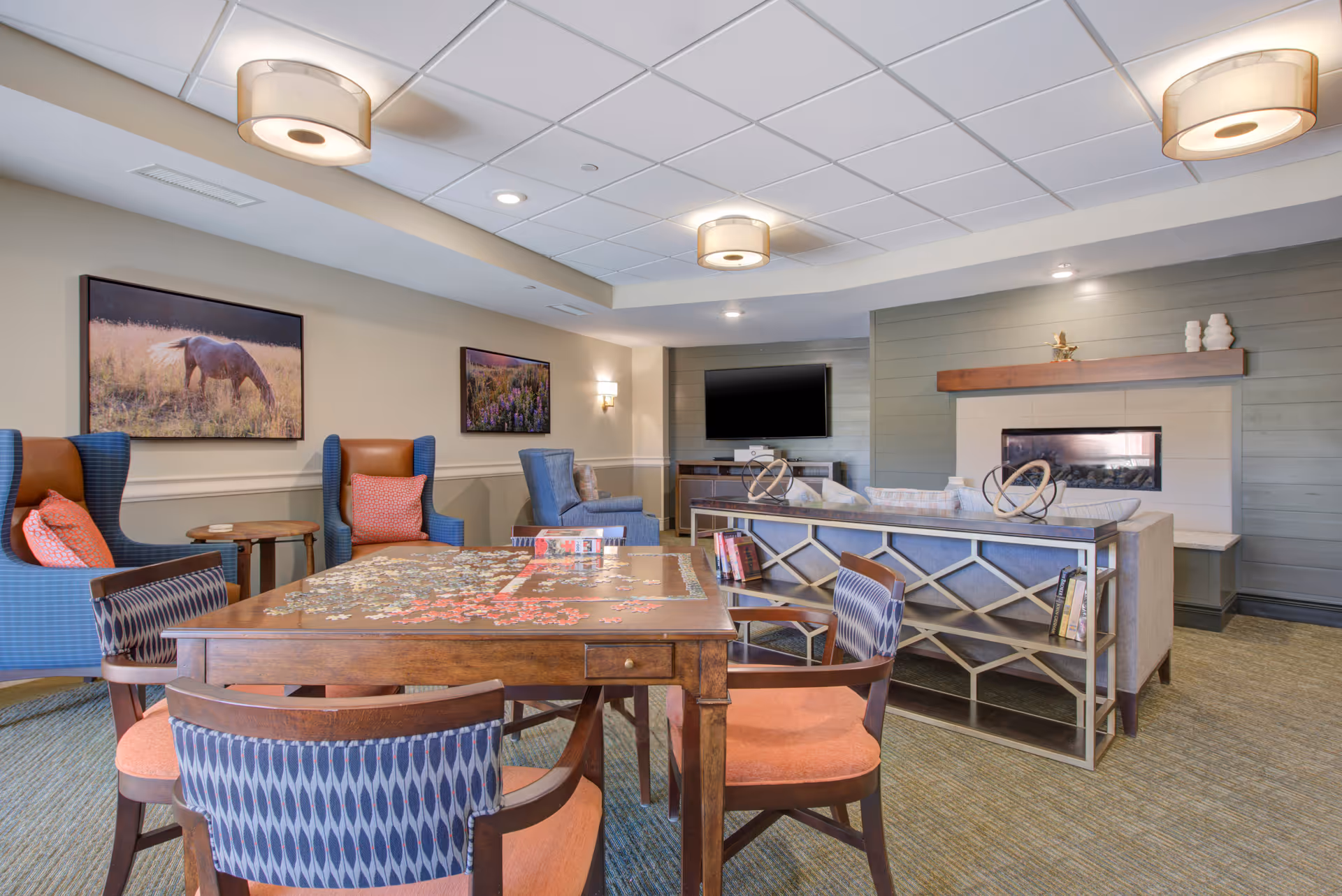 A cozy living room area in a senior living facility featuring a wooden table with a partially completed jigsaw puzzle, surrounded by four chairs with patterned upholstery. In the background, there are two blue armchairs with orange cushions, a beige sofa with a decorative metal back, a flat-screen TV mounted on the wall, and a modern fireplace with a wooden mantel. The walls are decorated with framed pictures of nature scenes, and the ceiling has recessed lighting and two round ceiling lights.