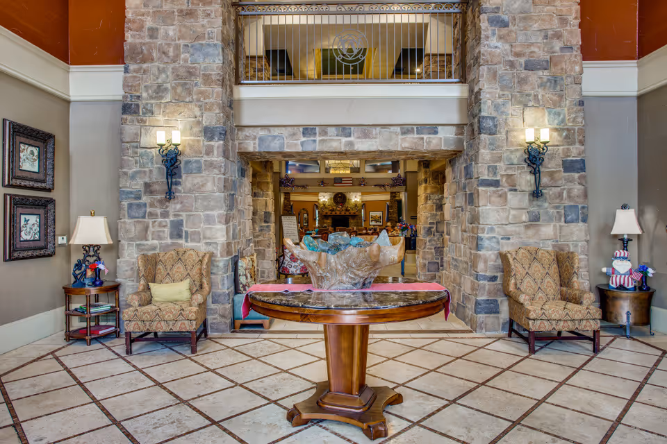 A spacious and elegant interior lobby area with stone walls and a tiled floor. In the center, there is a round wooden table with a decorative centerpiece. On either side of the table, there are two patterned armchairs with side tables and lamps. Wall sconces provide lighting on the stone pillars. The background shows an open doorway leading to another room with more seating and a fireplace.