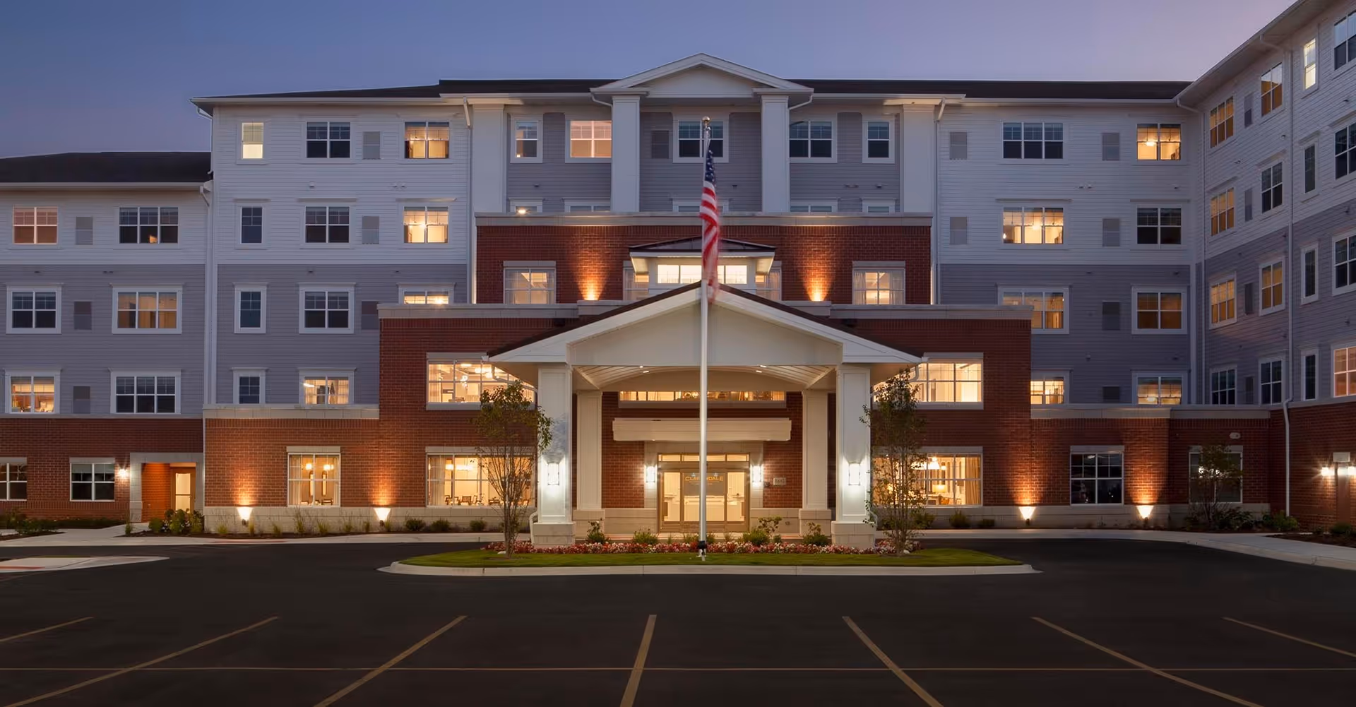 Front exterior view of Clarendale of Addison senior living facility at dusk, showing a large multi-story building with lit windows, a covered entrance with columns, an American flag on a flagpole, and a parking area in front.