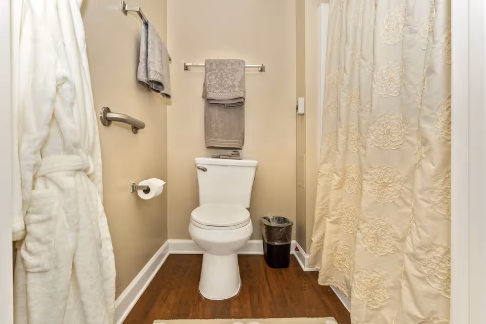Compact bathroom featuring a toilet centered between a towel rack and a shower with a decorative curtain, plus a robe and grab bar.