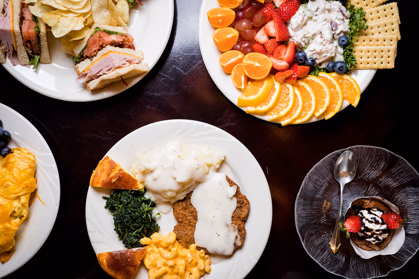 Top-down view of several plates on a dark table holding sandwiches, sliced fruit, mashed potatoes with gravy, greens, mac and cheese, and a dessert.