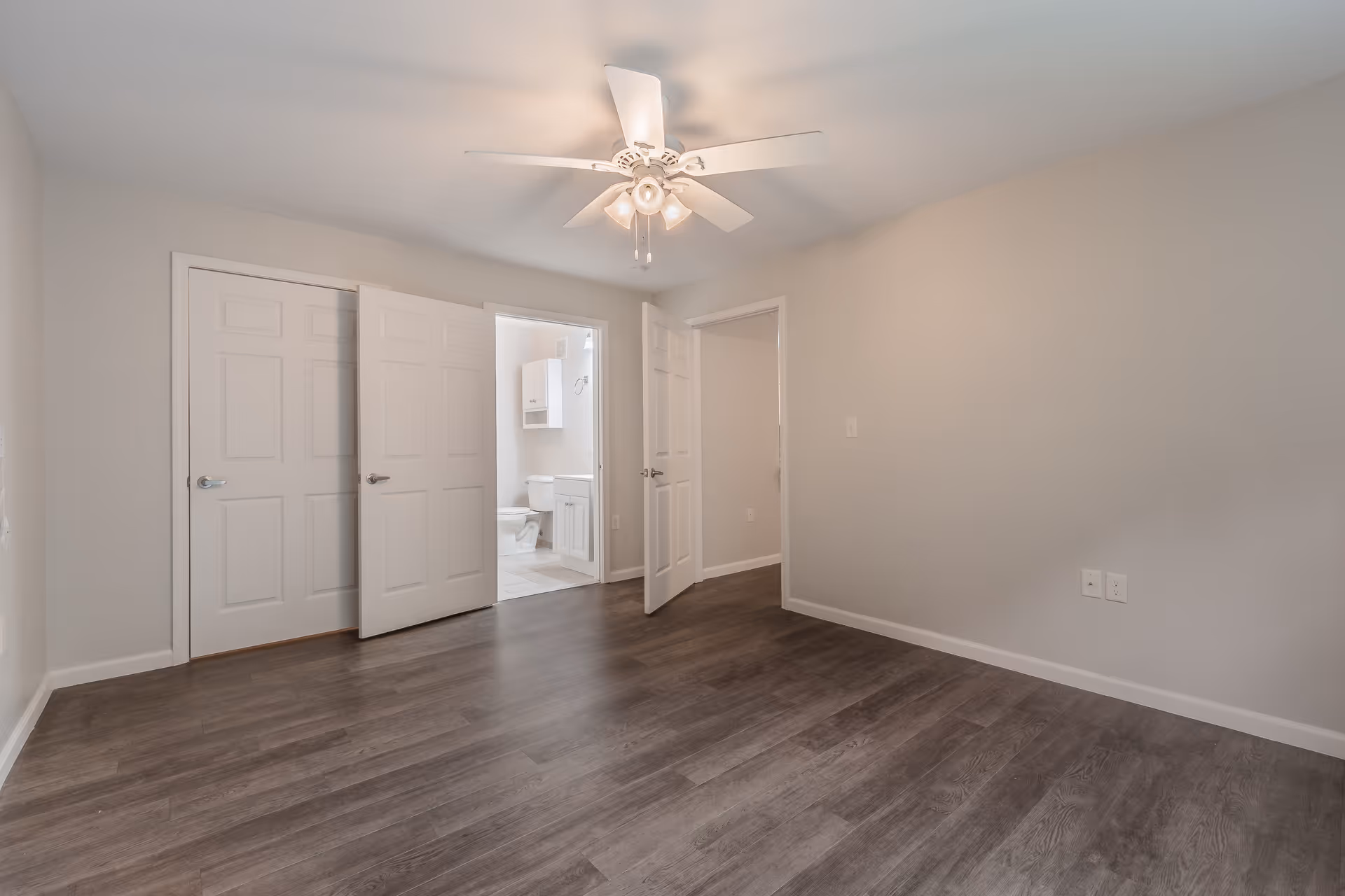 Empty room with gray wood flooring, light gray walls, and a white ceiling fan with lights. Two doors are open, one leading to a bathroom with a toilet and vanity visible, and the other to another room. There is a closed closet door on the left side.