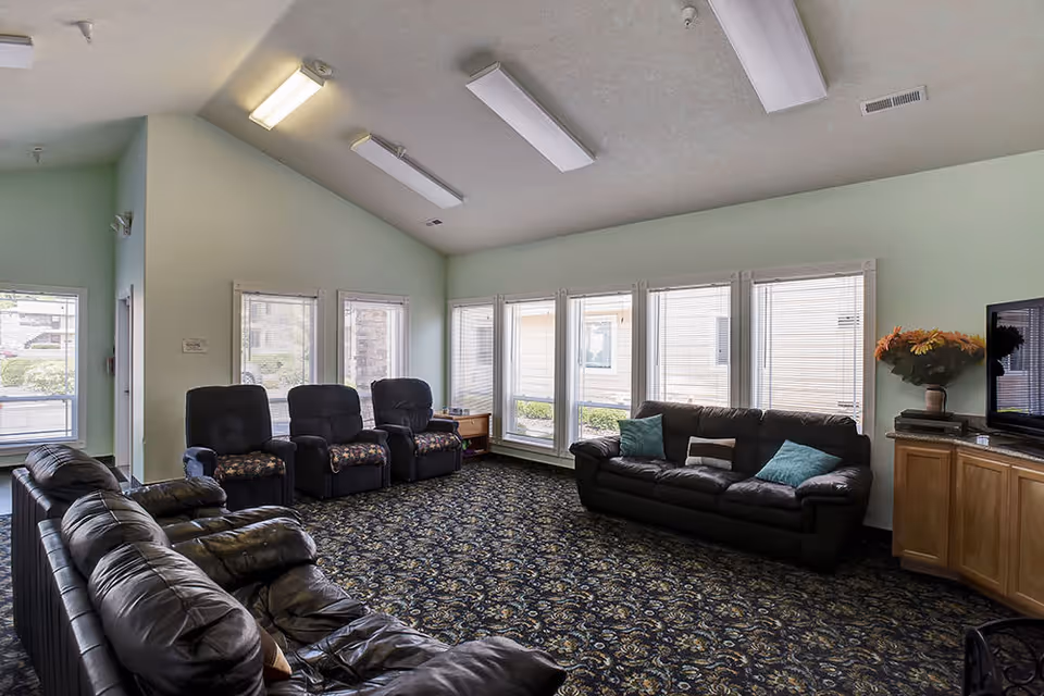 A spacious living room with multiple black leather sofas and recliners arranged around a patterned carpet. The room has large windows with blinds, light green walls, a high ceiling with fluorescent lights, and a wooden cabinet with a TV and a vase of flowers on top.