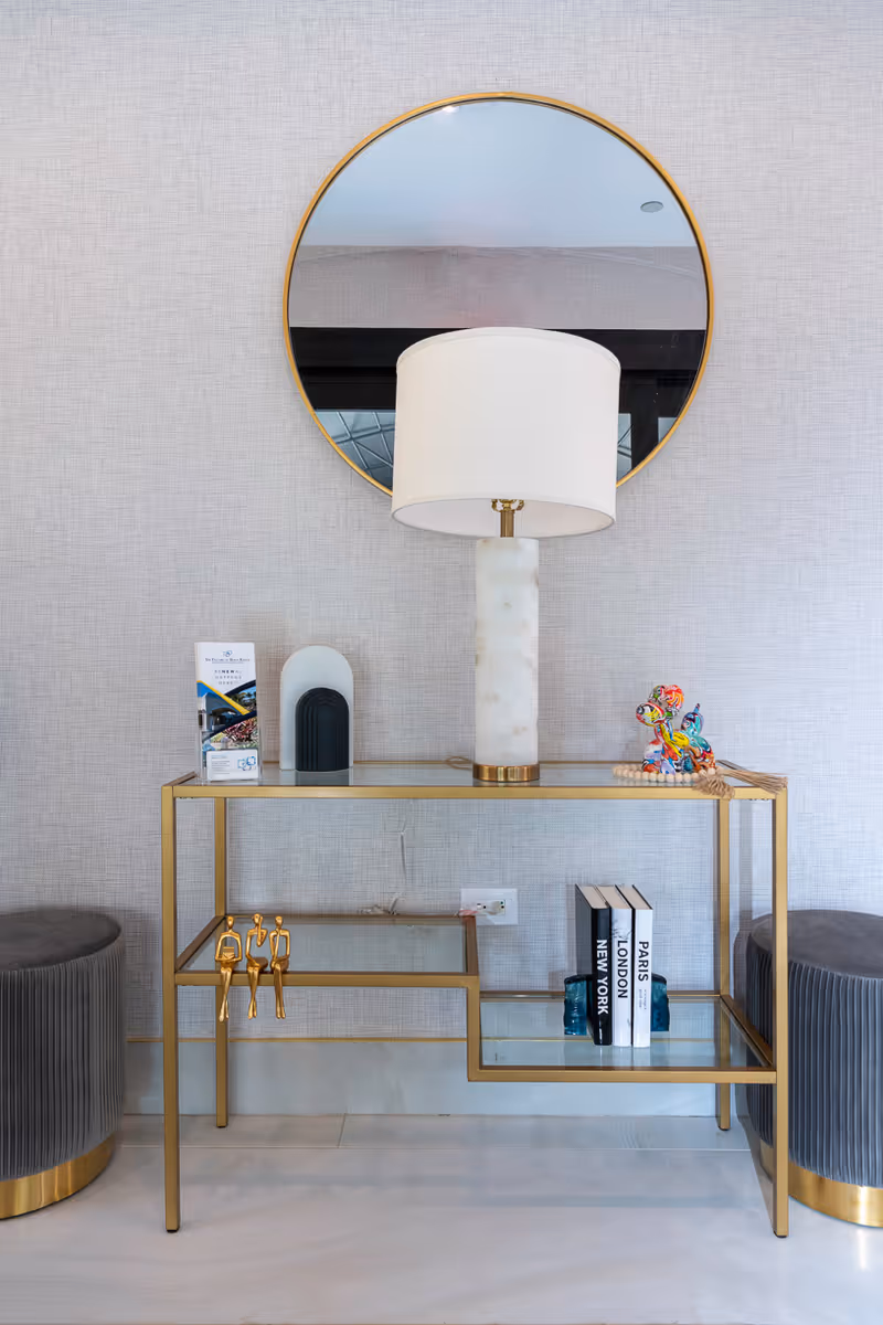 A modern interior setup featuring a gold-framed glass console table with decorative items including a white marble lamp with a white shade, a colorful decorative figurine, three books titled New York, London, and Paris, and two small blue vases. Above the table is a round mirror with a gold frame. On either side of the table are two gray upholstered stools with gold bases. The background wall is light gray with a subtle texture.